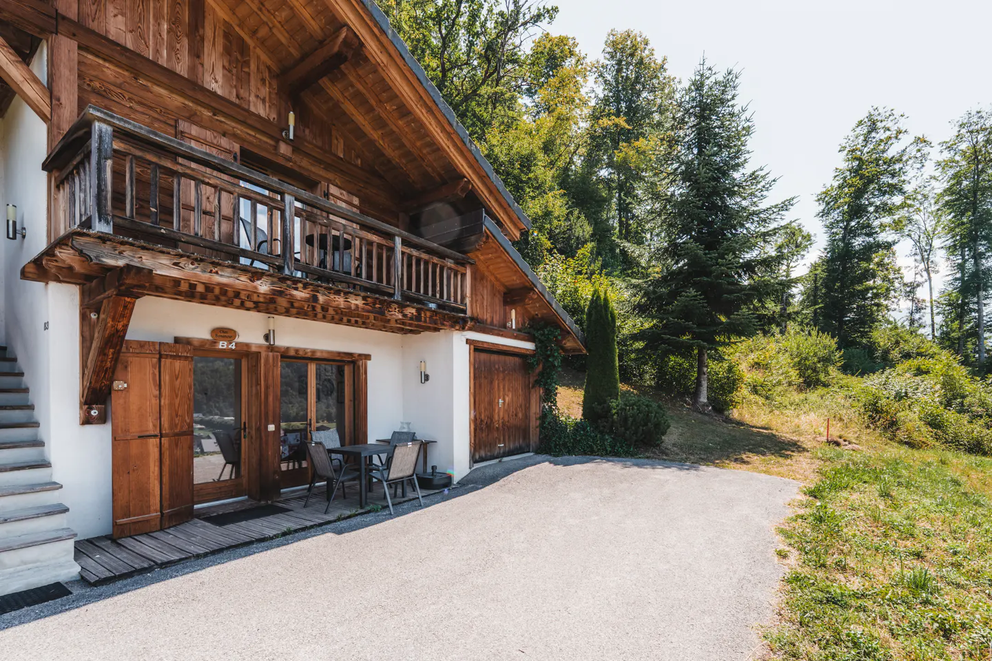 A two-story chalet with a wood balcony and a table with chairs outside. The chalet is surrounded by trees.