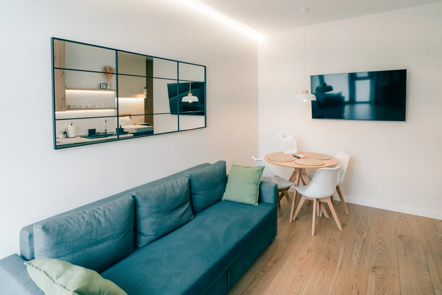 A bright living room with a blue sofa, green pillow, round table with white chairs, and a large mirror reflecting the kitchen.