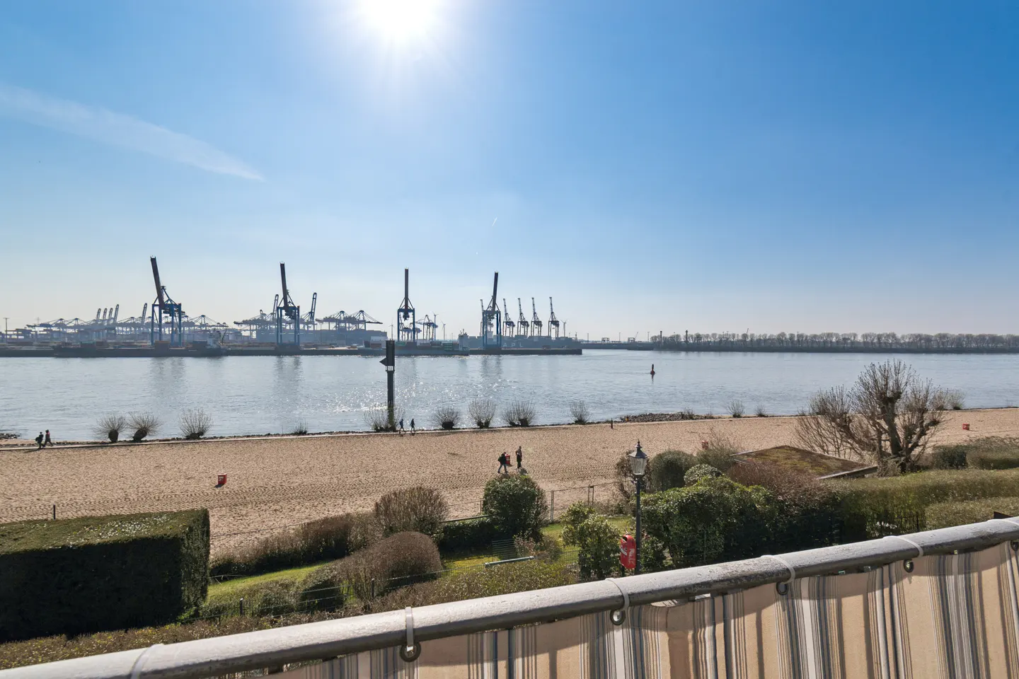 View from a balcony overlooking a sandy beach, a river, and a port with cranes under a sunny blue sky.