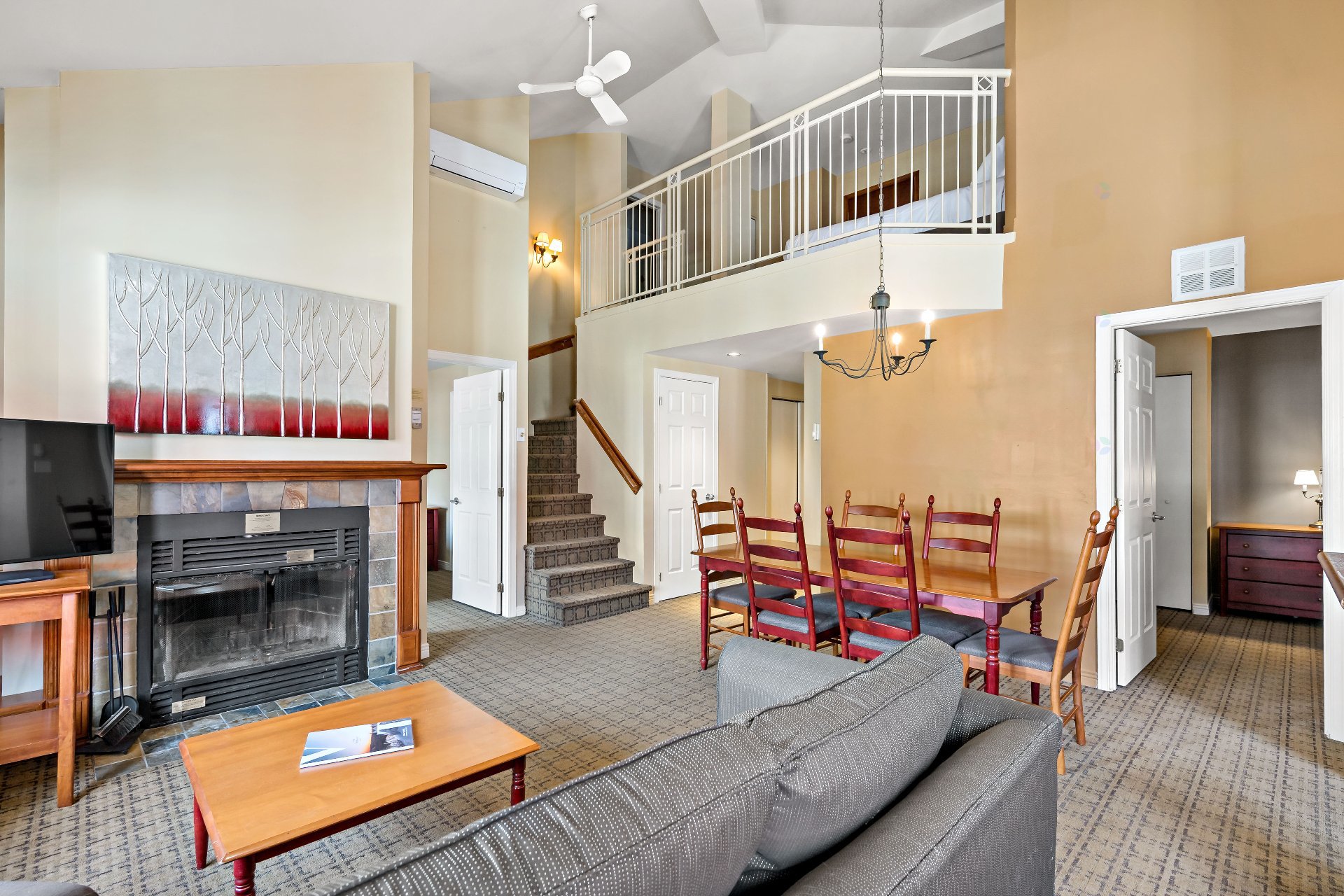 A living room with a fireplace, a gray sofa, a dining table with red chairs, and a staircase leading to a loft.