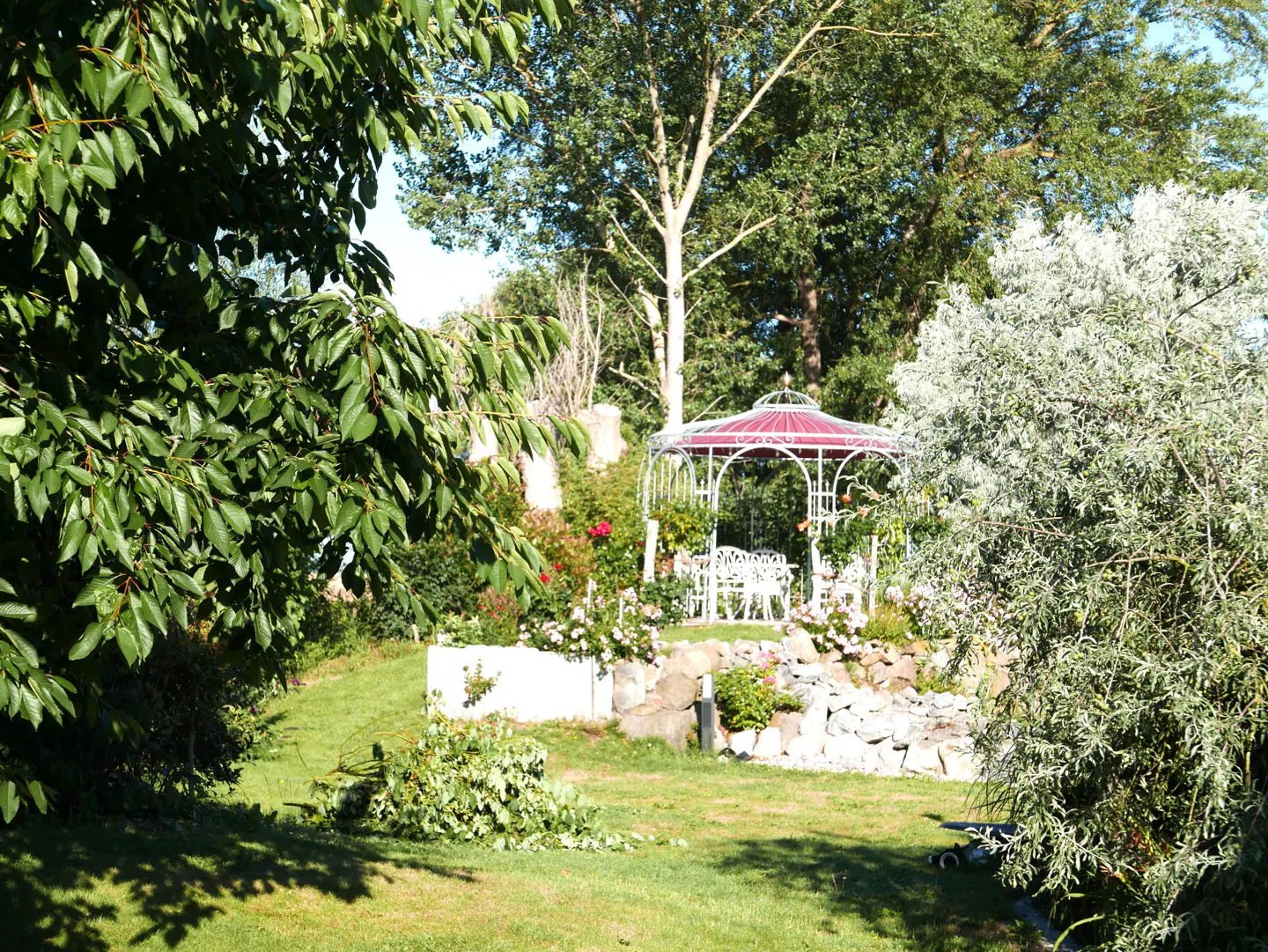 A white gazebo with a red roof sits in a lush green garden with trees and flowers.