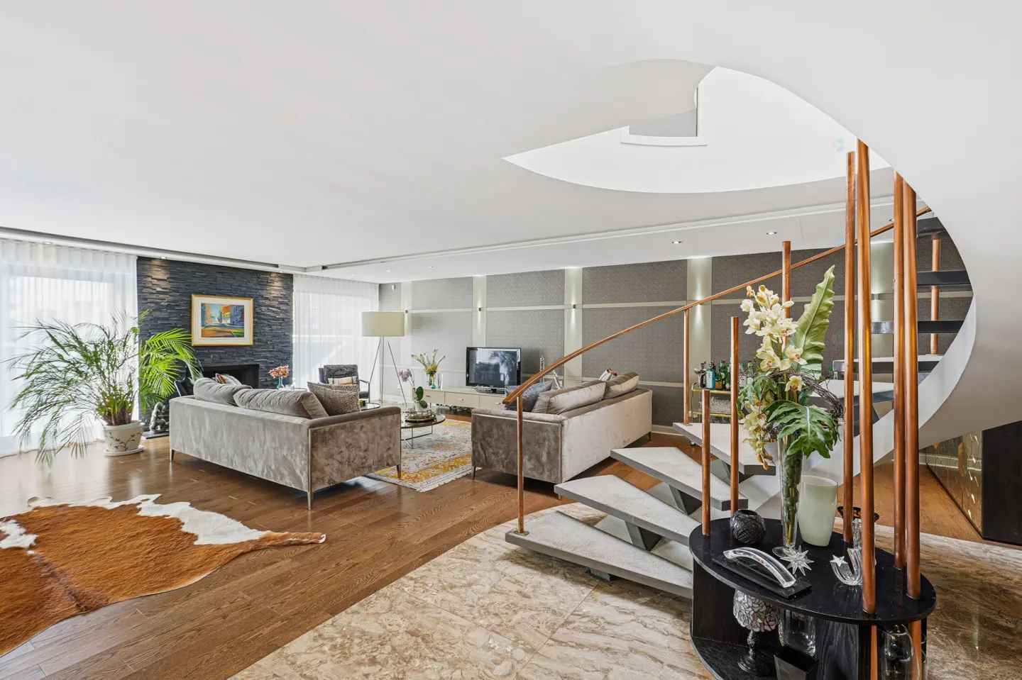 Bright living room with hardwood floors, grey sofas, a stone fireplace, and a spiral staircase with copper railings.
