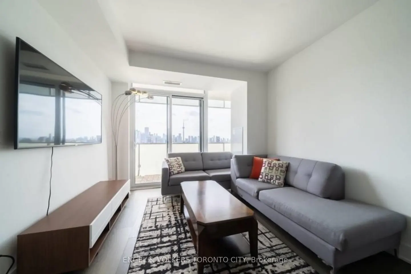 Modern living room with grey sectional sofa, wooden coffee table, wall-mounted TV, and a balcony view of the Toronto skyline.