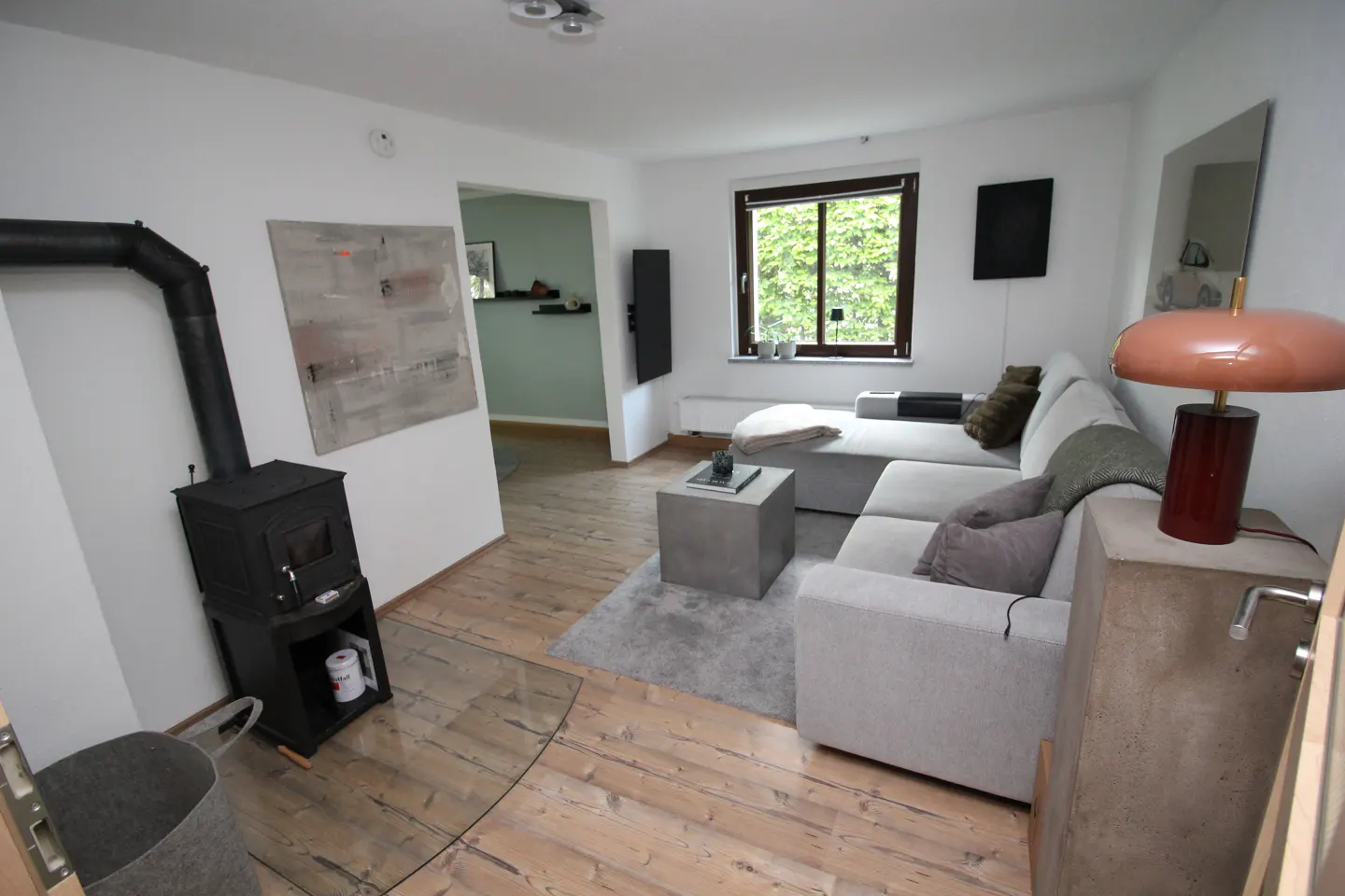 A living room with a wood-burning stove, a gray sofa, and a concrete side table with a lamp. A window is visible in the background.