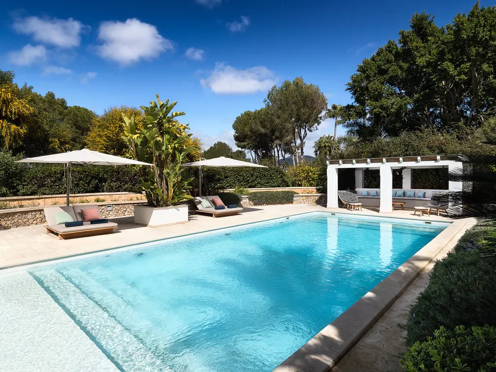 A bright blue swimming pool is surrounded by lounge chairs, umbrellas, and lush greenery. A white pergola with seating is in the background.