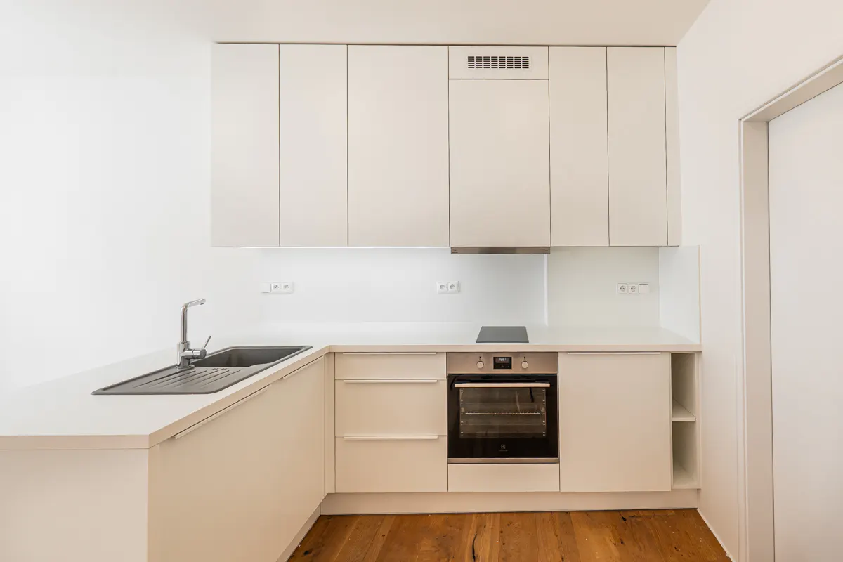 A modern, minimalist kitchen with white cabinets, countertops, and walls. A stainless steel sink and oven are visible. The floor is made of wood.