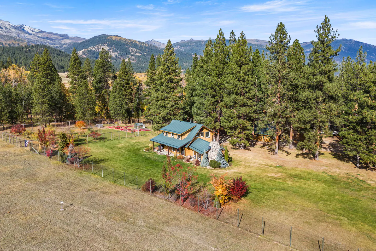 Aerial view of a yellow house with a green roof, surrounded by tall trees and mountains in the background.