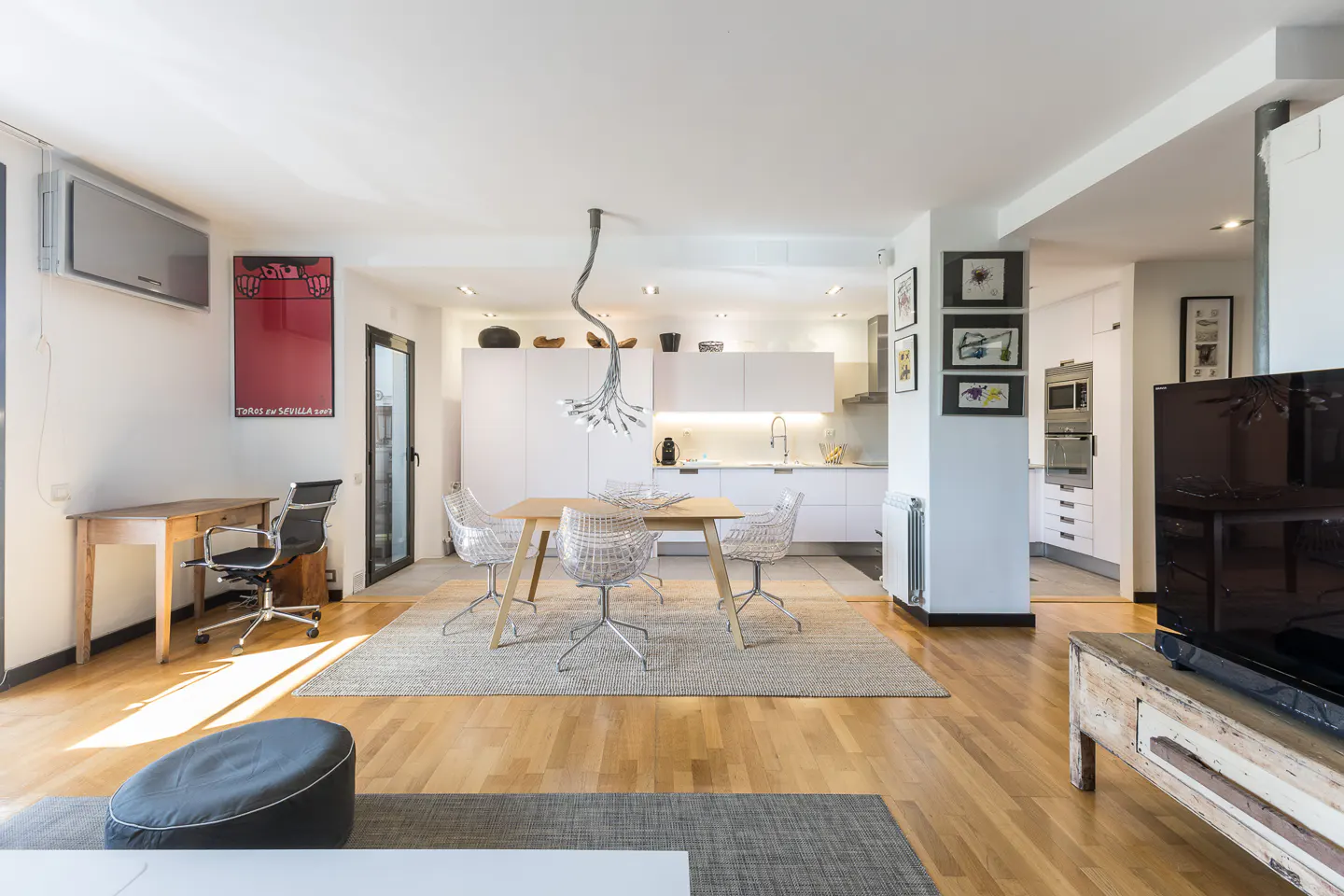 Bright, open-concept living space with wood floors, a dining table with clear chairs, and a modern white kitchen in the background.