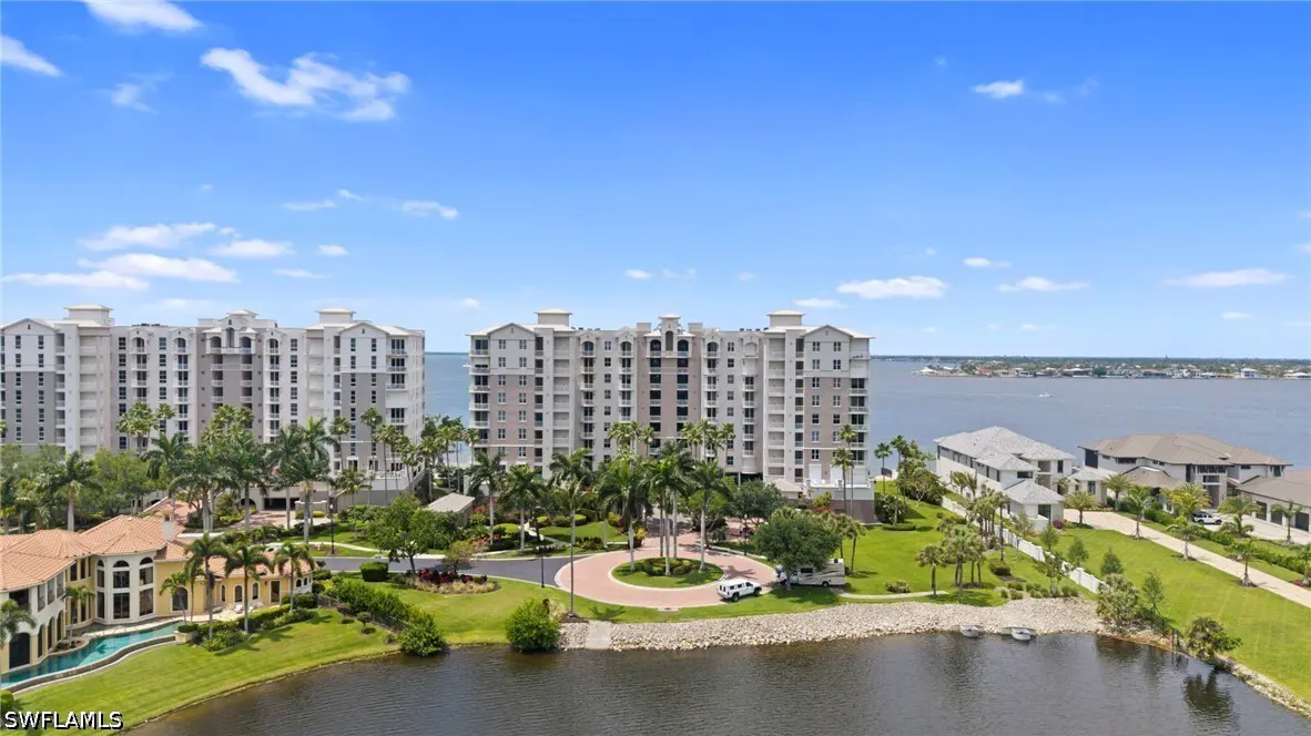 Aerial view of waterfront luxury condos with palm trees, a circular driveway, and a pool under a bright blue sky.