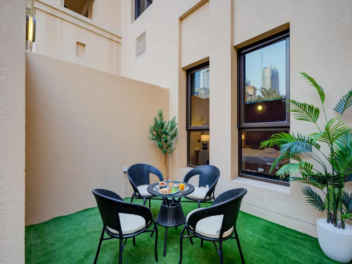 A small patio with artificial grass, four black chairs with white cushions, and a round table set for breakfast.