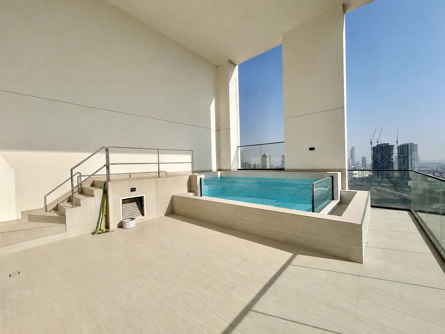 Modern rooftop terrace with a glass-walled plunge pool, beige tiled flooring, and a city skyline view under a clear sky.