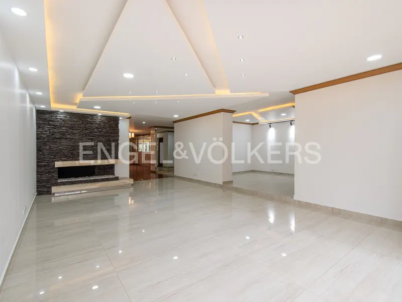 A bright, empty living room with white walls, tile floors, and a stone fireplace. Recessed lighting and the Engel & Völkers logo are visible.
