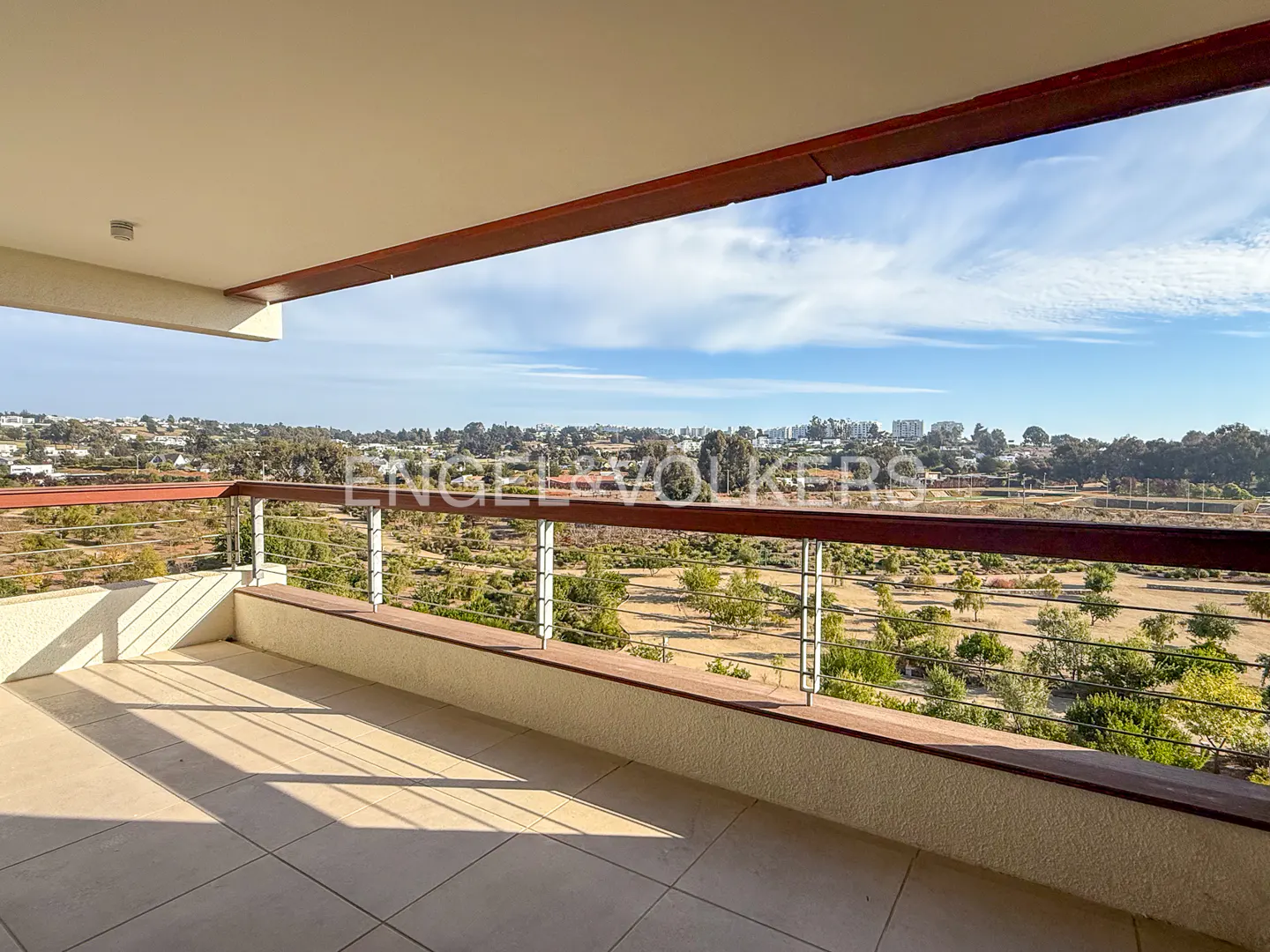 Balcony view with tile floor, metal railing, and wood trim. Distant trees and buildings under a blue sky.