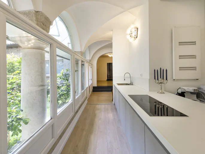Bright, modern kitchen with arched windows and a view of greenery. White countertops, gray cabinets, and a sleek cooktop. A hallway leads to another room.