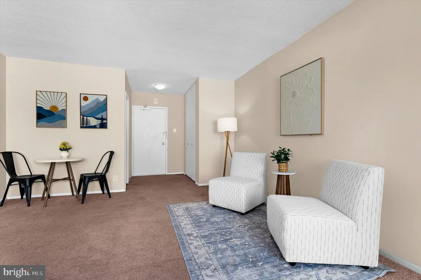 A living room with beige walls, brown carpet, and a blue rug. Two white chairs face each other, and a small table with black chairs sits nearby.