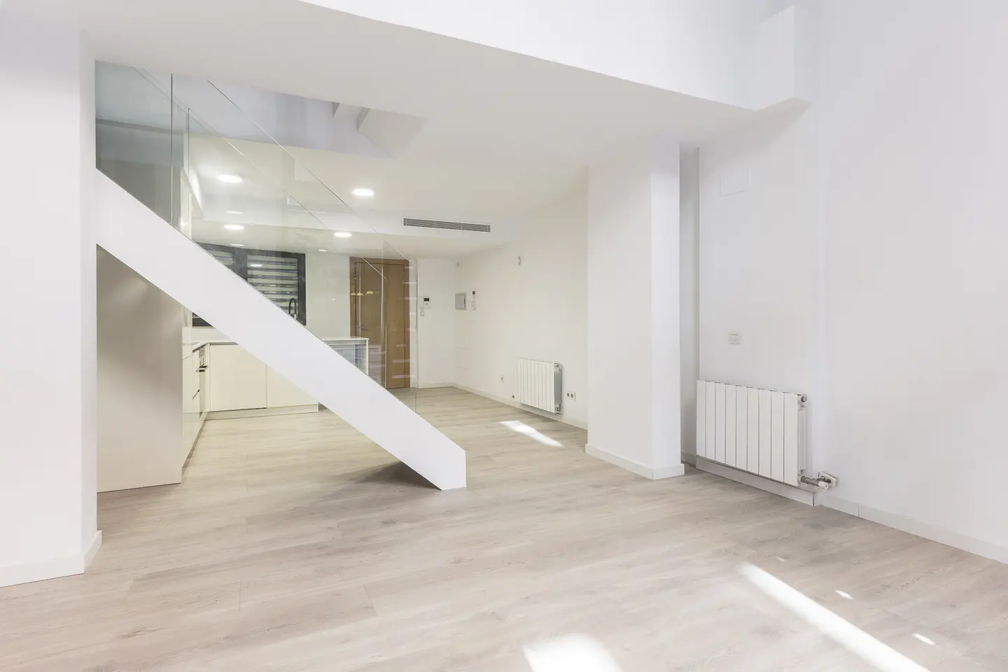 Bright, modern interior with white walls, light wood floors, and a white staircase with glass railing. A kitchen is visible in the background.