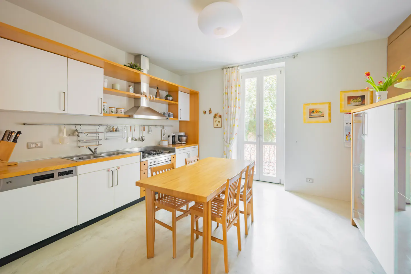 Bright kitchen with white cabinets, orange trim, and a wooden table with chairs. A window with patterned curtains lets in natural light.