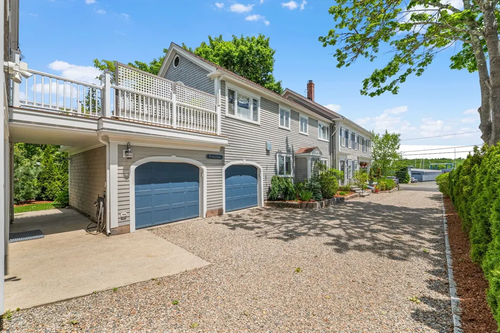 Gray house with blue garage doors, a gravel driveway, and a second-story deck under a bright blue sky.
