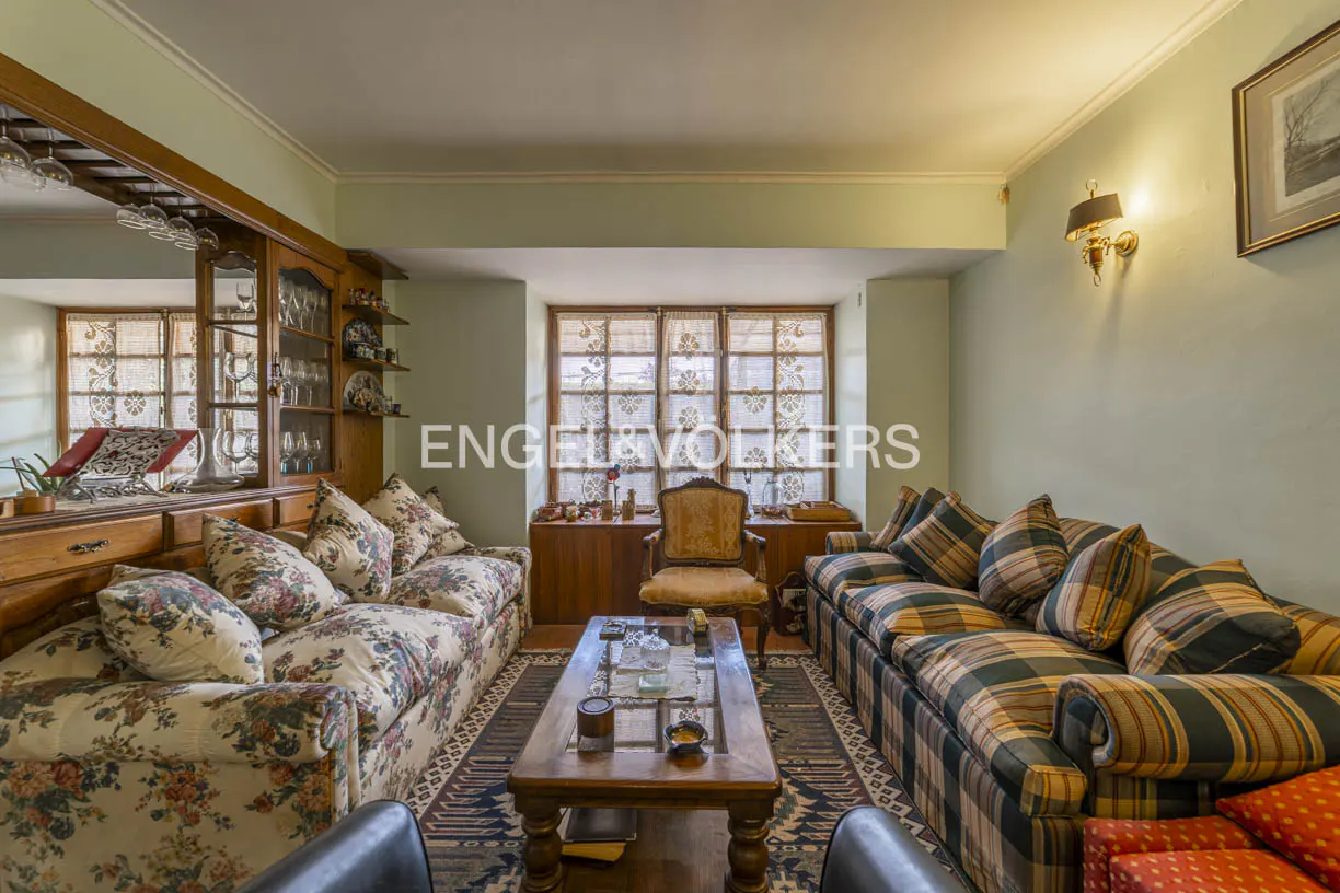 Living room with floral sofa, plaid sofa, wood coffee table, and built-in wood shelving with glassware. Pale green walls.