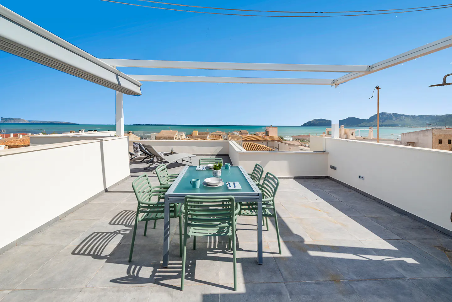 Rooftop patio with a table set for six, green chairs, and ocean view. White pergola overhead, blue sky.