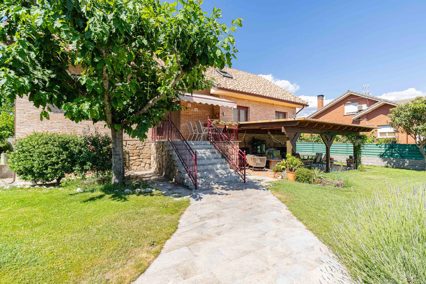 Stone steps lead to a brick house with a covered patio, green lawn, and a large tree under a clear blue sky.