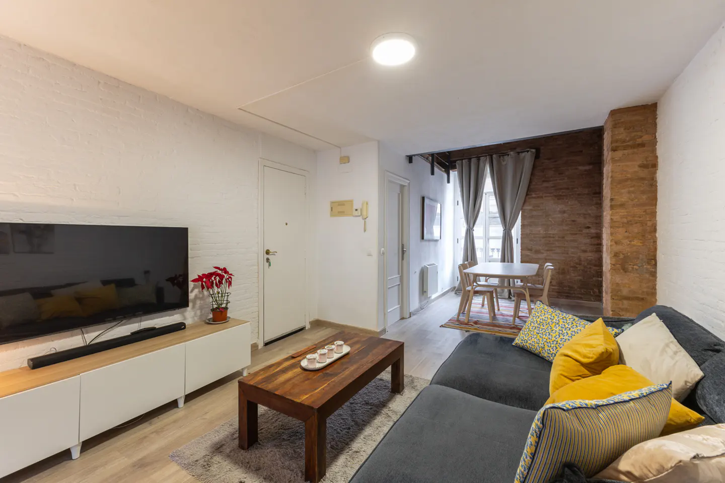 Living room with gray sofa, wooden coffee table, and TV on a white cabinet. Dining table near a brick wall with gray curtains.