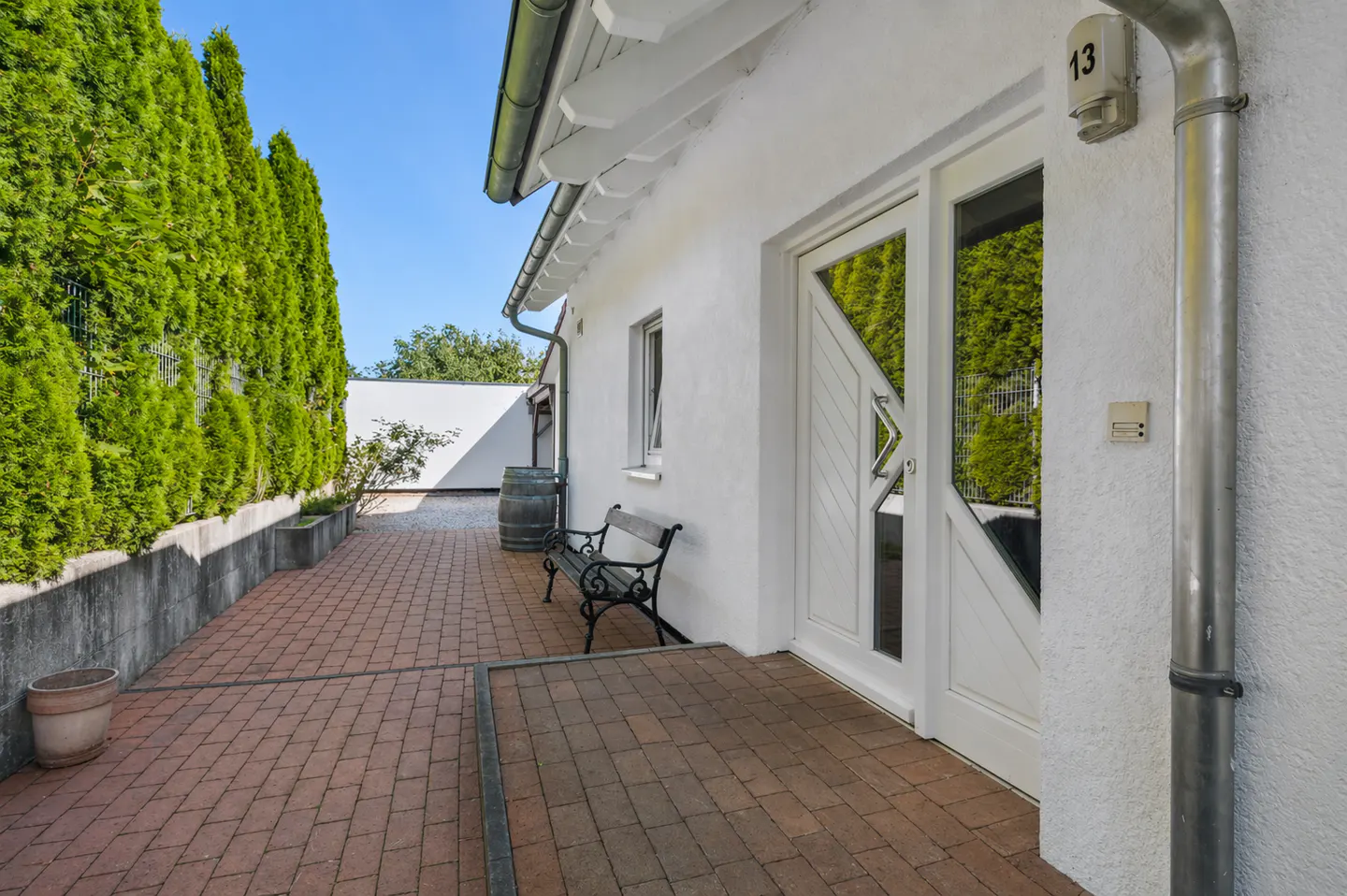 Exterior view of a white house with a brick patio, green hedges, and a white door with the number 13 above it.