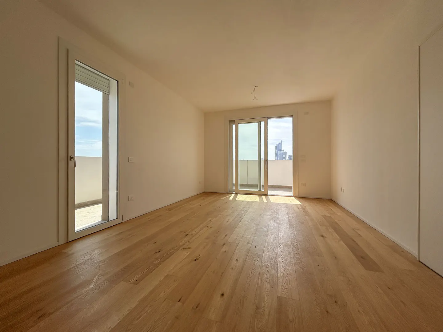 Bright, empty room with light wood floors, white walls, and balcony access. Sunlight streams in through sliding glass doors.