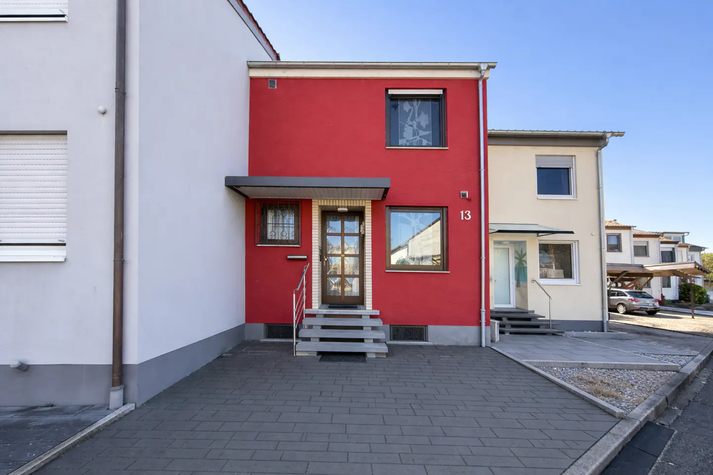 Exterior view of a two-story red house with a gray roof, steps, and a door with glass panels. The house number "13" is visible on the wall.