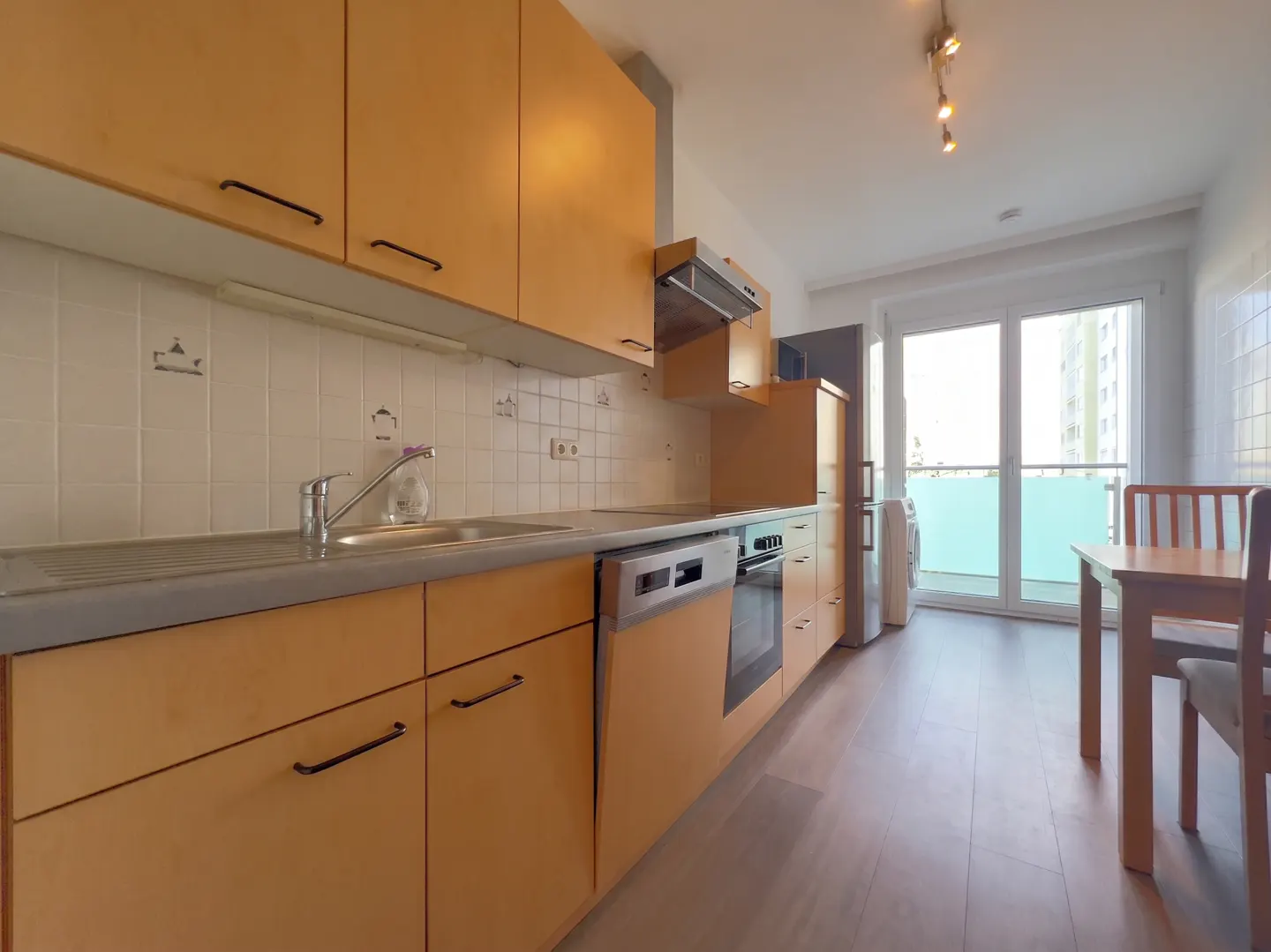 A bright kitchen with light wood cabinets, a gray countertop, and a stainless steel sink. A table and chairs are near a balcony with a view of a tall building.