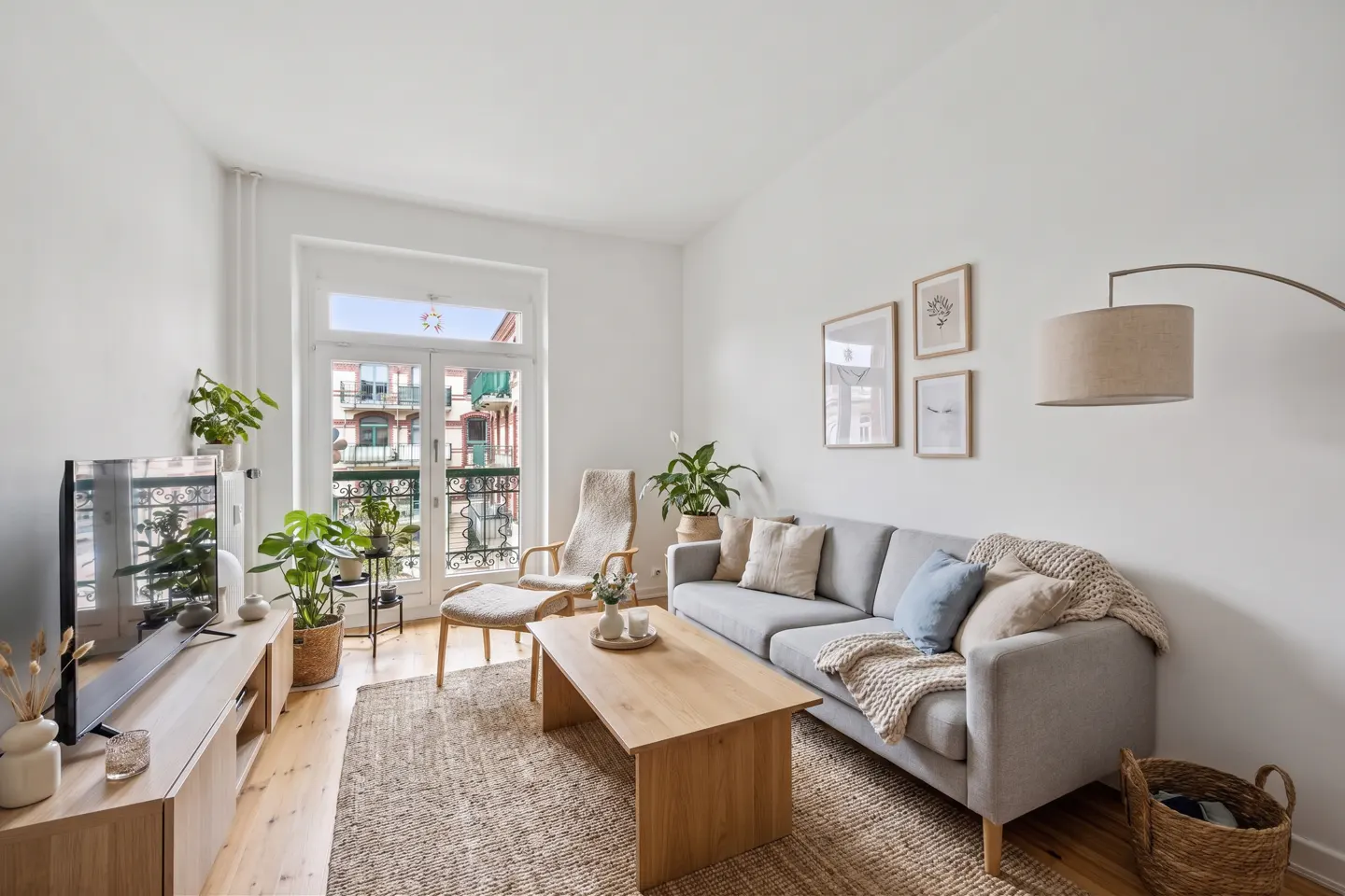 Bright living room with a gray sofa, wooden coffee table, and a chair near a balcony door. Plants and framed art add a touch of nature.