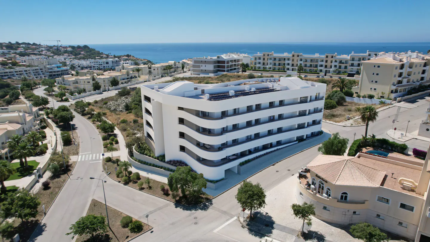 Aerial view of a modern white apartment building with curved balconies, located near the coast in a sunny residential area.