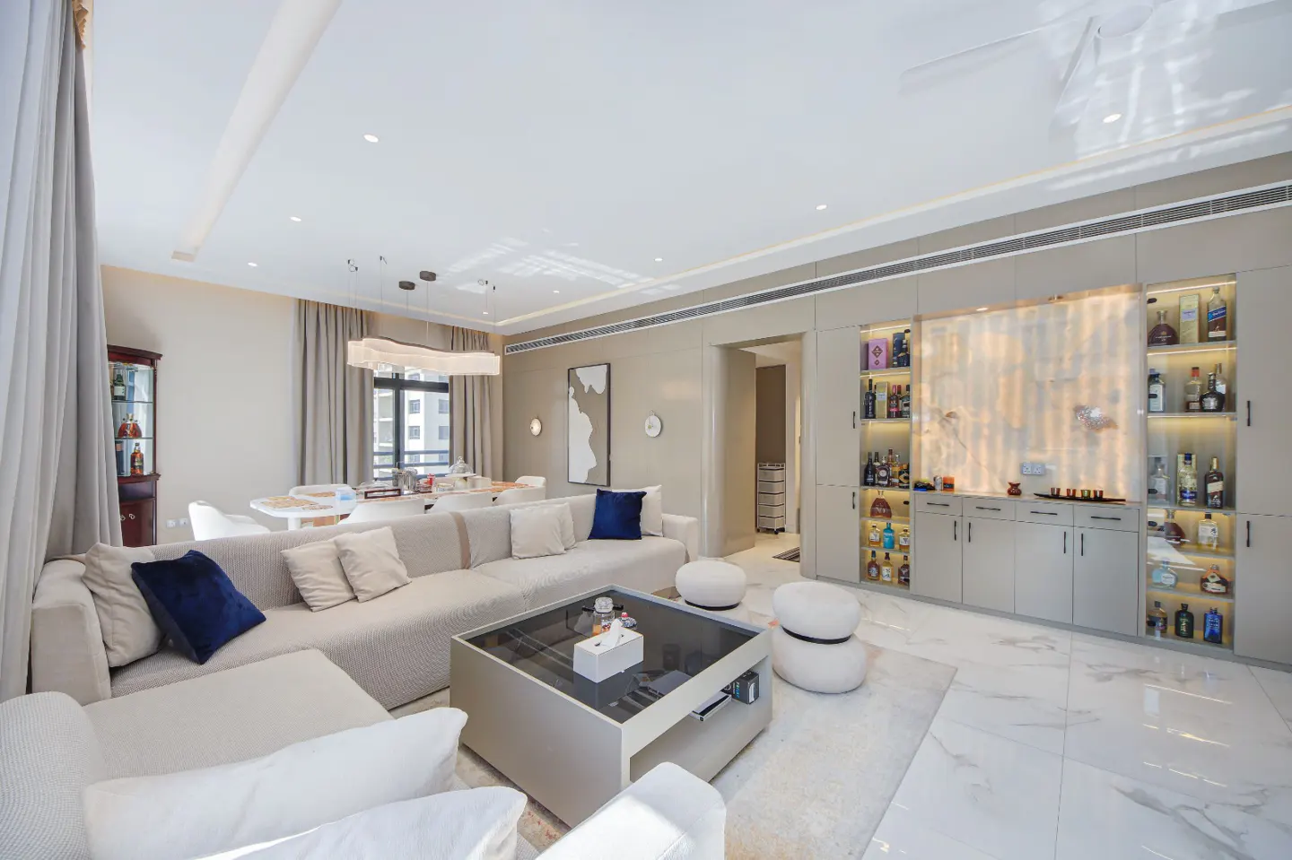 Bright living room with a large beige sectional sofa, glass coffee table, and built-in bar with liquor bottles.