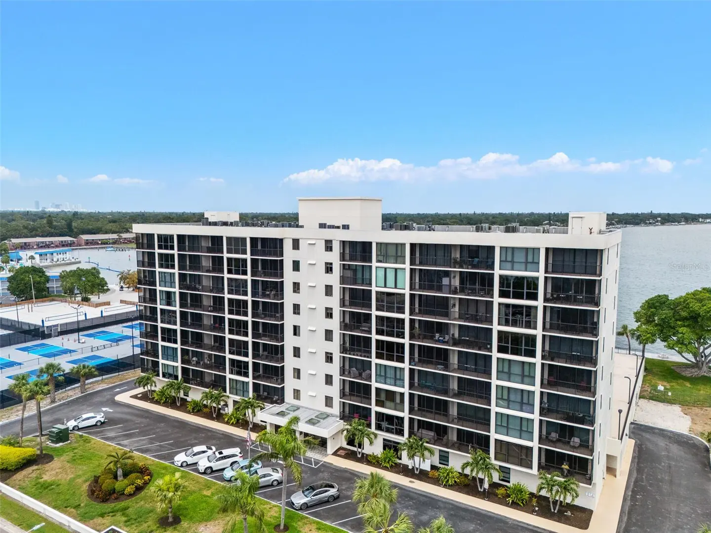 Aerial view of a white, multi-story condo building with black balconies, a parking lot, and palm trees under a blue sky.