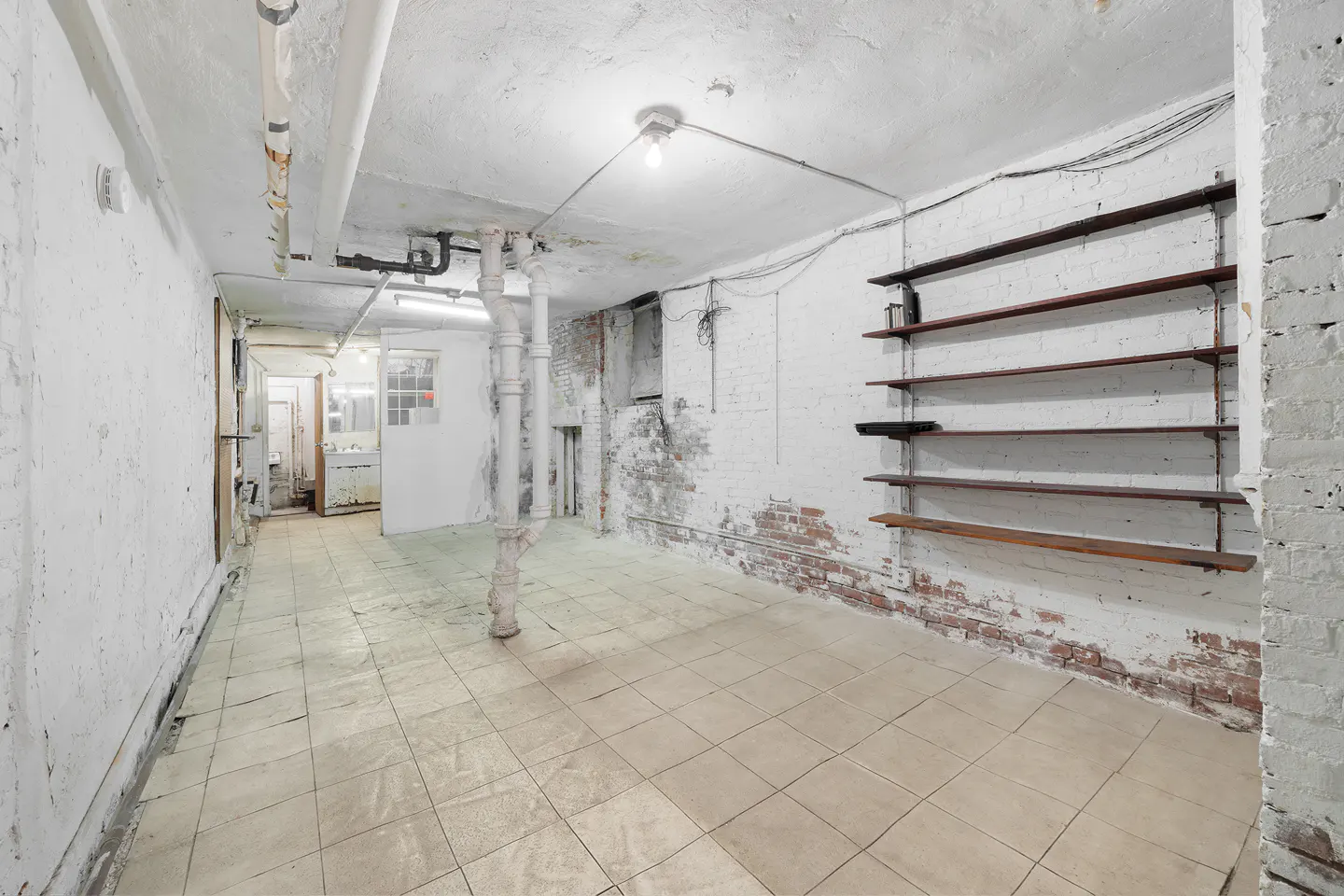 A basement with white-painted brick walls, tile floors, and wooden shelves. Pipes run along the ceiling.
