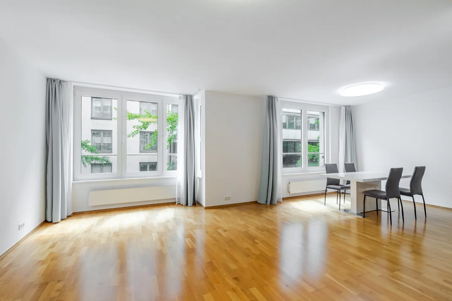 Bright, empty room with hardwood floors, white walls, and large windows with gray curtains. A white table and four chairs sit in the corner.