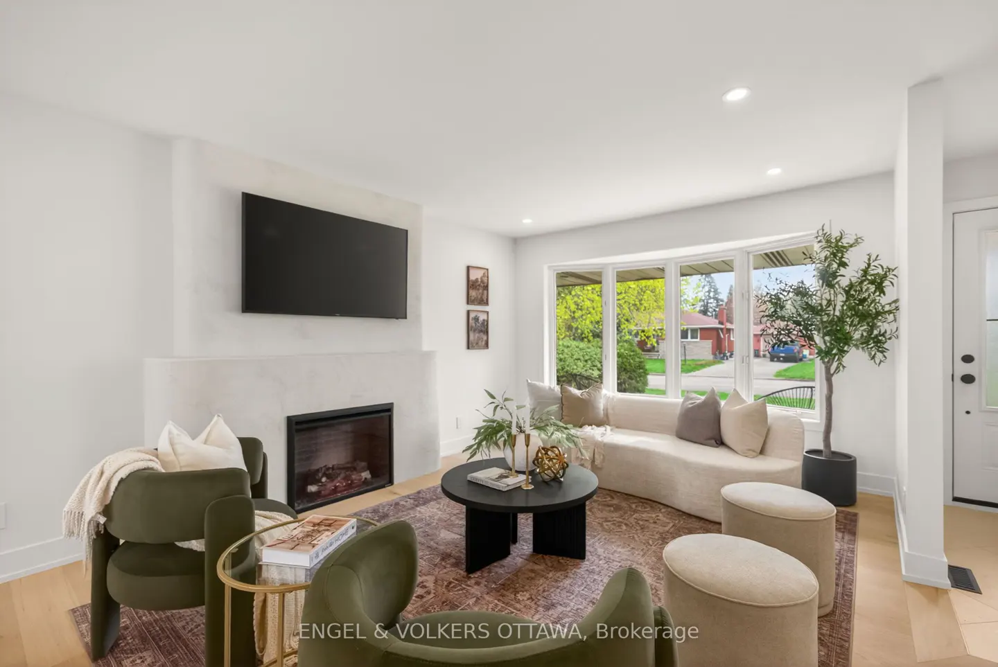 Bright living room with fireplace, TV, and bay window. Olive green chairs, beige sofa, and black coffee table on a patterned rug.