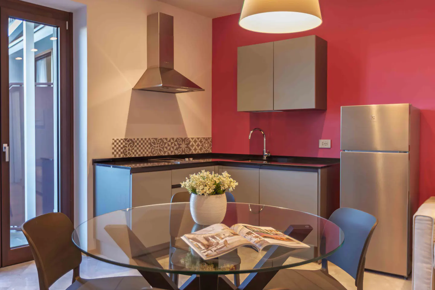 A modern kitchen with a glass table, chairs, and stainless steel appliances. The walls are painted in pink and white.