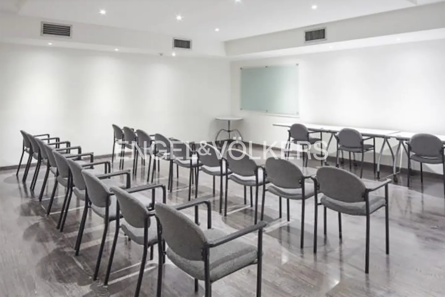 A bright, empty conference room with rows of gray chairs facing a whiteboard and tables.