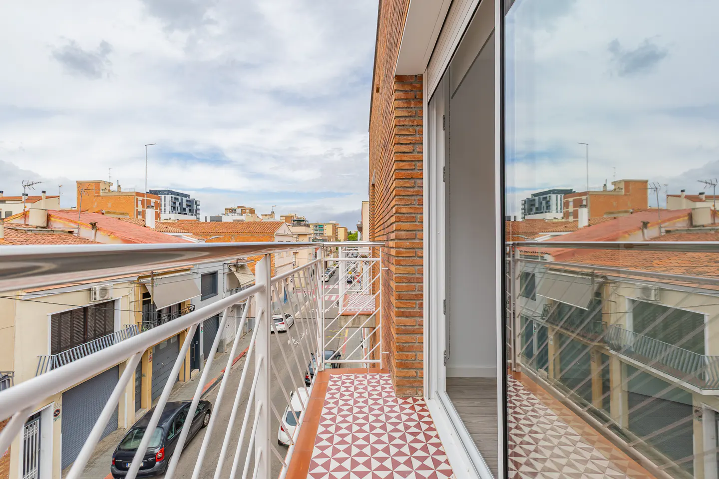 Modern balcony with red and white patterned tile floor, white metal railing, and a view of a city street below.