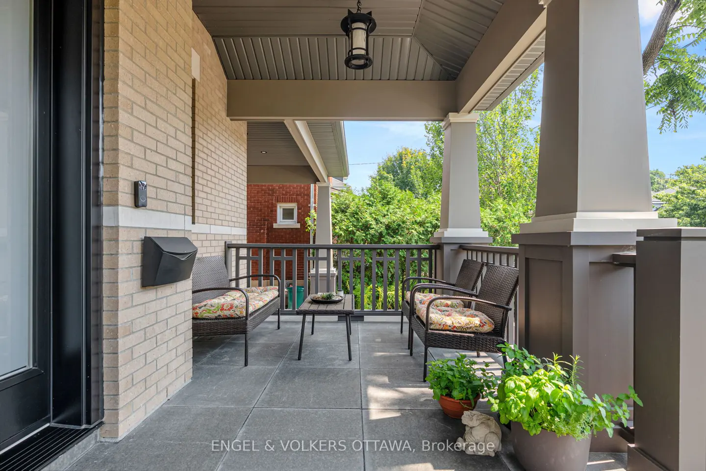 Covered porch with two wicker chairs, a small table, and potted plants. Brick wall and large gray columns.