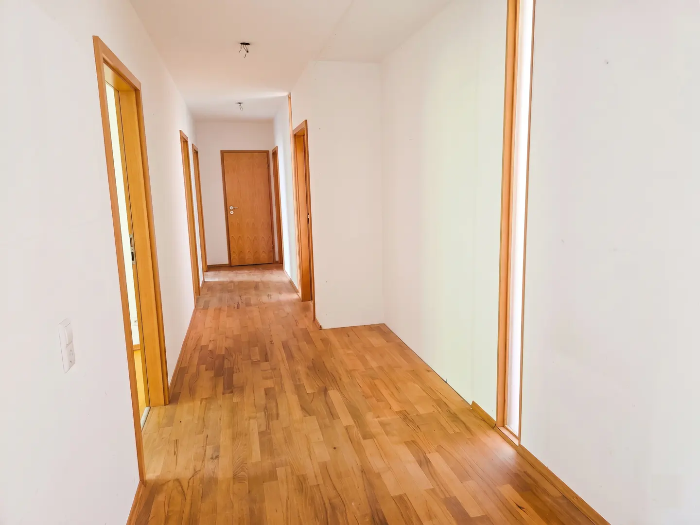 Bright hallway with white walls, light wood flooring, and multiple wooden door frames leading to various rooms.