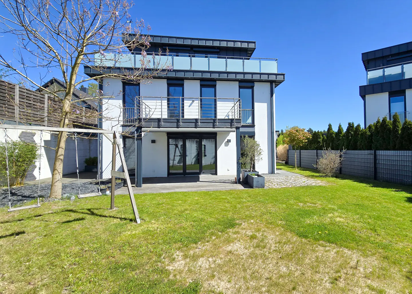 Modern two-story white house with black trim, balconies, and a green lawn. A swing set stands in the yard under a tree.