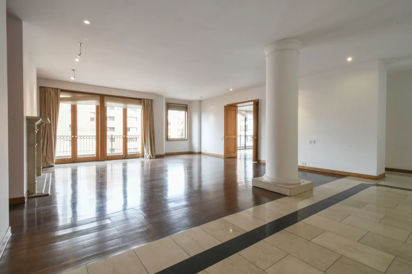 Bright, empty living room with hardwood floors, white walls, and large windows with tan curtains. A white column stands near an open doorway.