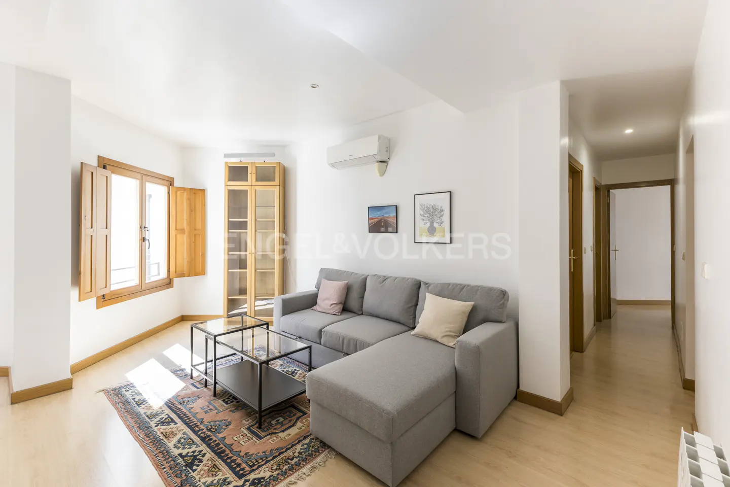 Bright living room with gray sectional sofa, glass coffee table, and patterned rug. Wooden cabinet and window with open shutters.