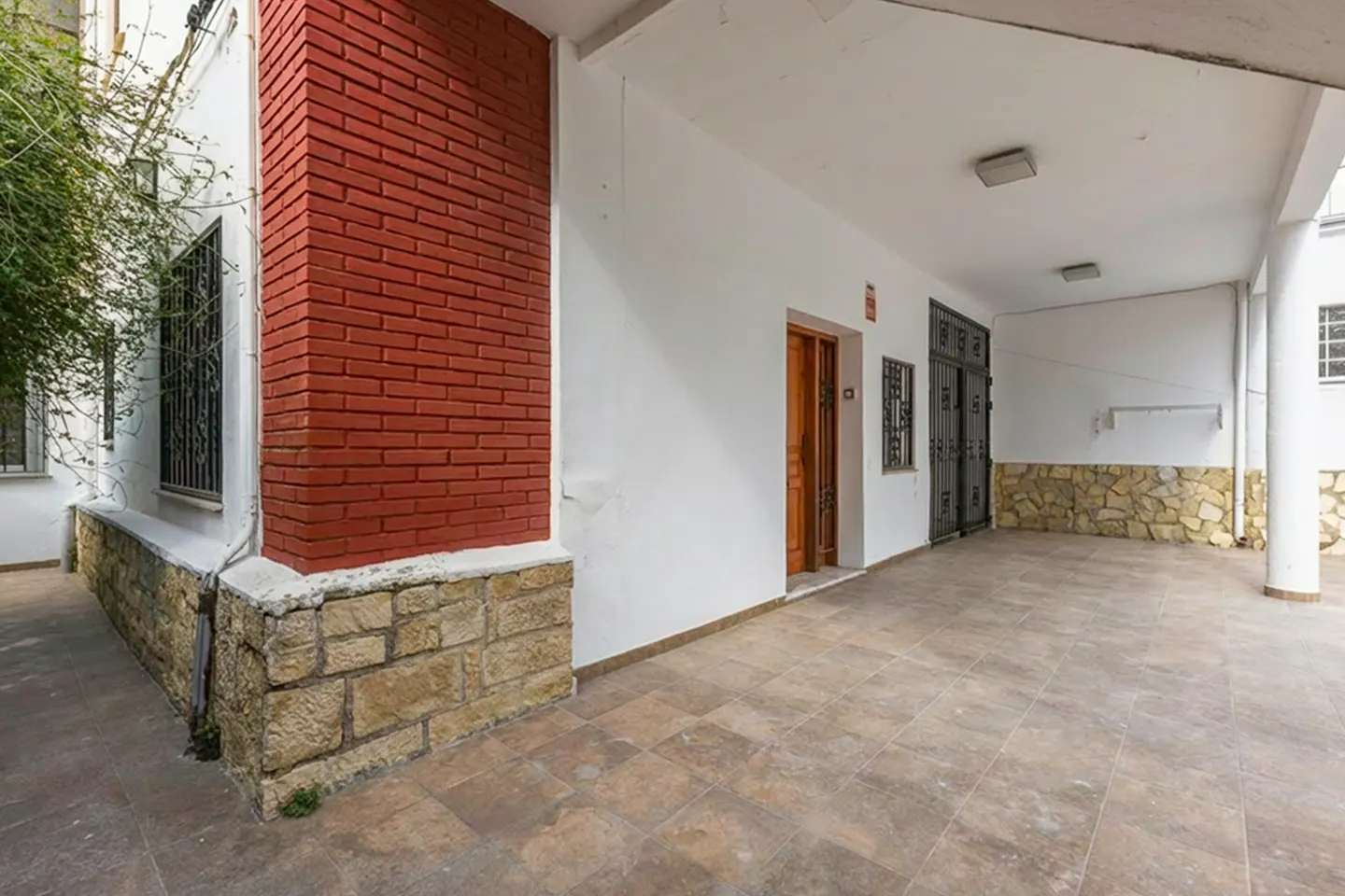 Covered patio with red brick corner, stone base, and tile floor. White walls with a wooden door and black iron gate.