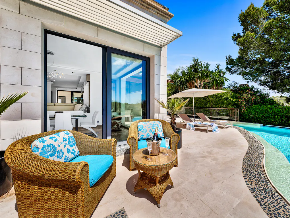 Outdoor patio with wicker furniture, blue cushions, and a bottle of champagne. A pool and lounge chairs are in the background.