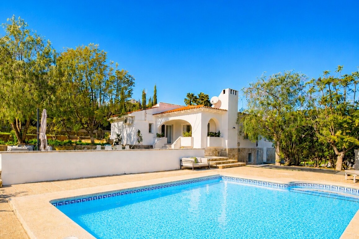 White villa with a terracotta roof and a bright blue swimming pool in the foreground, surrounded by trees under a clear sky.