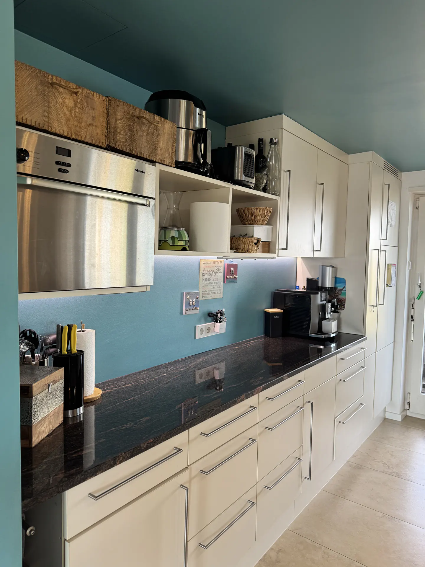 A modern kitchen with white cabinets, dark granite countertops, and a teal backsplash. Stainless steel appliances are visible.