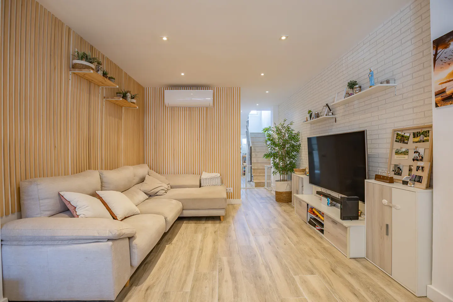 Living room with a beige sectional sofa, wood-look floors, and a large TV. The walls are paneled and brick, with shelves and plants.